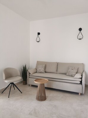 Ein minimalistisches Wohnzimmer mit einer Couch, einem Sessel, einem kleinen Beistelltisch und einer Topfpflanze.