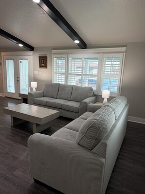 Ein modernes Wohnzimmer mit einem grauen Sofa, einem Couchtisch und einer Lampe. Es gibt Fenster mit Jalousien, eine Tür und ein Bild an der Wand.