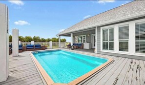 Ein Schwimmbad mit Holzterrasse, eine überdachte Terrasse mit Sitzgelegenheit und ein Haus mit weißem Zierleistenwerk und Fensterläden.