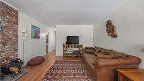 A living room with a brick wall, a television on a wooden stand, a brown leather sofa, and a patterned rug. - Riverkern Retreat House (Kernville)