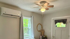 Ein Zimmer mit Deckenventilator, Klimaanlage, einer weißen Tür, einem Fenster mit Vorhängen, einem weißen Sofa mit Kissen und einem kleinen Tisch mit Gegenständen darauf.