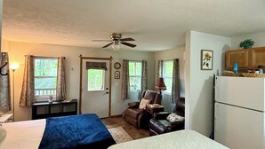 Ein gemütlicher Raum mit Bett, Esstisch, Stuhl, Deckenventilator, Kühlschrank und einem Fenster mit Vorhängen.