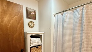 Ein Badezimmer mit Holztür, einem weißen Schrank mit Körben, einem waschraumtypischen Vorhang mit Muster und einer Wanduhr.