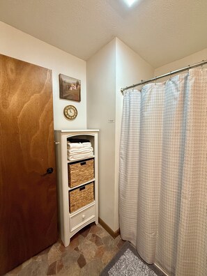 Een badkamer met een houten deur, een wit kastje met manden, een douchegordijn met een patroon en een wandklok.