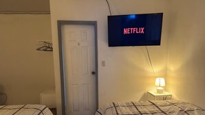 Ein Hotelzimmer mit zwei Betten, einem Fernseher, der 'NETFLIX' anzeigt, einer Tür und einem Nachttisch mit einer Lampe.