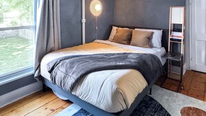 A bedroom with a bed, a bedside table, a lamp, a window with curtains, and a view of the outdoors. - The Quaint House: Modern Heritage in Roundout Kingston Waterfront (Kingston)