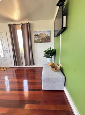 Une pièce avec un plancher en bois, un banc gris, un mur vert, un tableau, une plante et un miroir.
