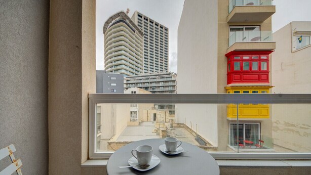 A balcony with a table and chairs, a window overlooking a cityscape with buildings. - 2BR Apt in St Julian’s Heart (St. Julian`s)