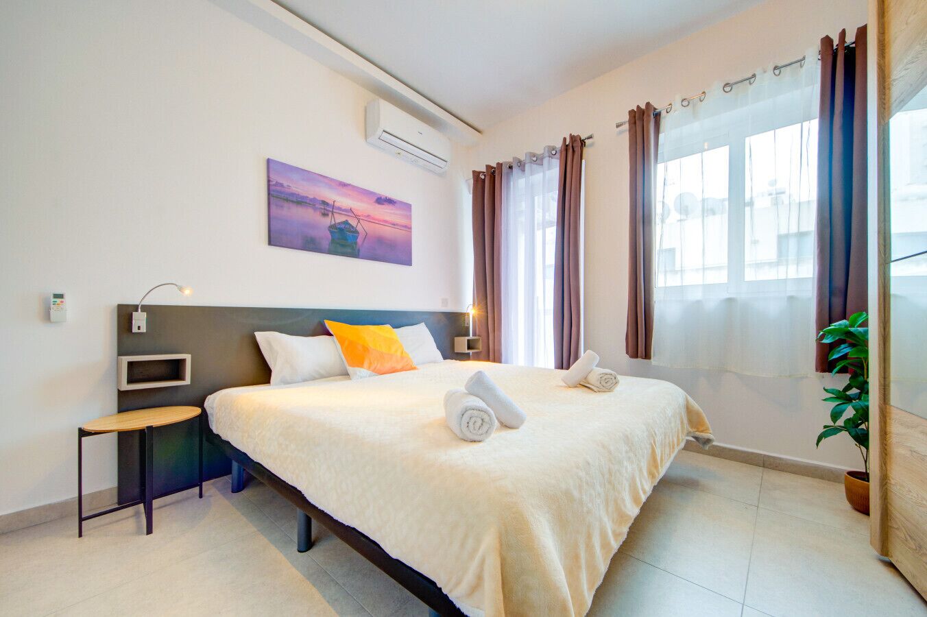 Ein Hotelzimmer mit Bett, Nachttisch, Klimaanlage, Fenster mit Vorhängen und einem Bild an der Wand.