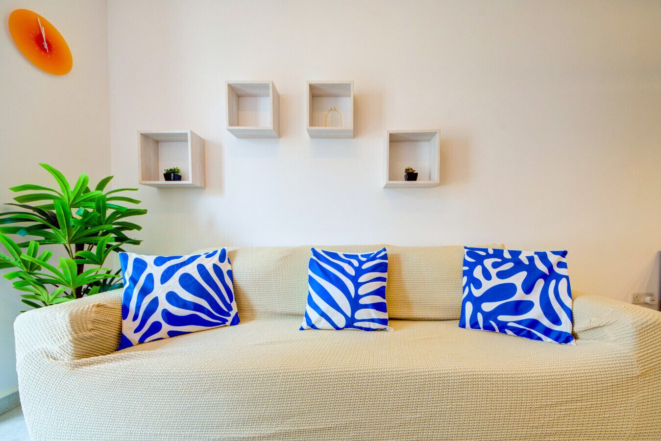 A living room with a beige sofa, three blue patterned cushions, a potted plant, and four white shelves with small items.