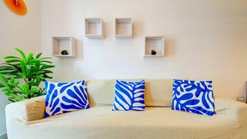 A living room with a beige sofa, three blue patterned cushions, a potted plant, and four white shelves with small items.