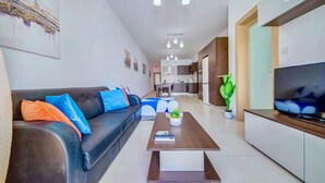 Ein modernes Wohnzimmer mit Sofa, Couchtisch und Fernseher. Im Hintergrund ist eine Küche zu sehen.
