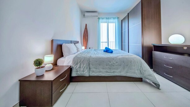 Ein modernes Schlafzimmer mit Bett, Nachttisch, Lampe und einem Fenster mit Vorhängen.