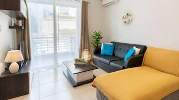 Un salon moderne avec un canapé, une table basse, une lampe, une plante et une vue dégagée sur l’extérieur grâce à une grande fenêtre.