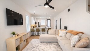 A modern living room with a sofa, a TV mounted on the wall, a ceiling fan, and a kitchen area in the background. - Modern 2BR Townhome - King + Queen - 20 Min to Downtown, IAH, & IAH - Gated (Houston)