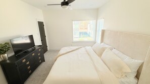 Un dormitorio con una cama, un televisor sobre un aparador, un ventilador de techo y una ventana con persianas.