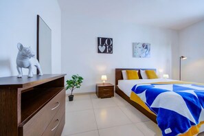 Ein Schlafzimmer mit Bett, Nachttisch, Kommode, Spiegel, einem Wandbild und einem Blumentopf.