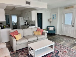 Ein Wohnzimmer mit einer Couch, einem Couchtisch, einer Küche im Hintergrund, einem Türkis-farbenen Sessel und einer Tür mit Fenster.