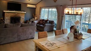 A cozy living room with a fireplace, a dining area with a table set for four, and a balcony with a view of trees. - Beach by the Lake -Cozy Stay with Gorgeous Views (Pocono lake)