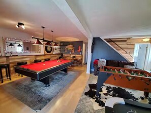 Mesa de billar, mesa de futbolín y zona de bar en un espacio con suelo de madera y una escalera.