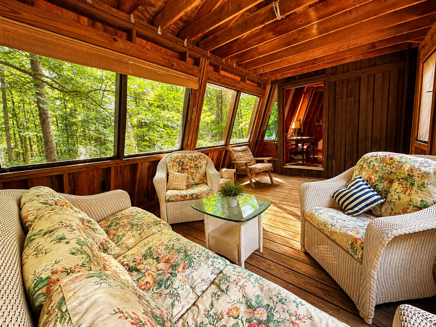 Interior de una cabaña de madera con sofá, sillones, una mesa de vidrio y ventanas amplias con vistas a un bosque.