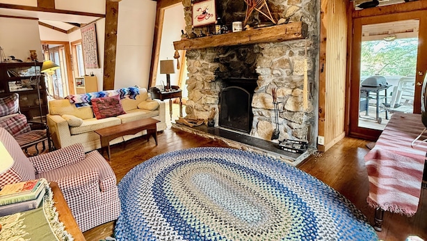A cozy living room with a stone fireplace, wooden beams, a round patterned rug, and a sofa with patterned cushions. - Rustic Pine Lodge - Cozy 4 BR Adirondack Escape (Schroon Lake)