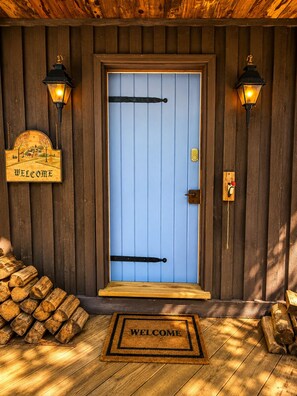 Une porte en bois avec un panneau bleu, deux lanternes fixées au mur, une pancarte de bienvenue, une sonnette et un paillasson.
