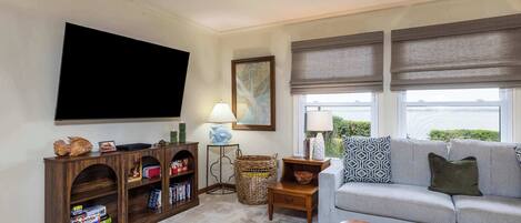 A living room with a flat-screen TV, a bookshelf, a lamp, a wicker basket, and a grey sofa with patterned cushions.