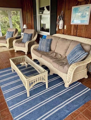 A wicker sofa set with cushions, a matching coffee table, and a blue striped rug on a wooden floor.