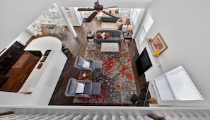 A spacious loft with a high ceiling, a dining area, a seating area with a red patterned rug, and a large window providing natural light.