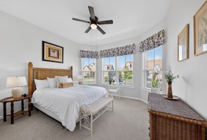 Une chambre à coucher comprenant un lit, des tables de chevet, un ventilateur de plafond, des fenêtres avec volets et une table d’appoint en osier.
