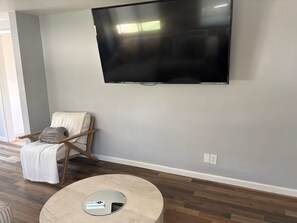Ein Wohnzimmer mit Flachbildfernseher an der Wand, einem Holzstuhl mit Kissen und einem runden Couchtisch mit Fernbedienung.