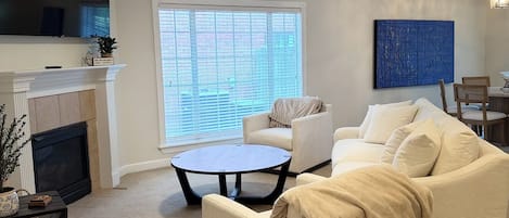 A living room with a fireplace, a ceiling fan, a large window with blinds, a white sofa, a coffee table, and a painting on the wall.