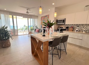 A modern kitchen with a central island, dining chairs, a ceiling fan, and a view of the ocean. - Oceanfront Corner Unit  Full Ocean View  Beachfront Pool Parking  Jaco (Jaco)