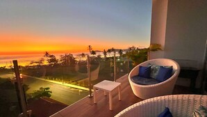 Un balcone con vista sul tramonto sull'oceano, una sedia in vimini con cuscini e un tavolino.