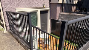 Un balcón con barandilla de metal negro, un pequeño jardín con terraza de madera y un edificio con pared exterior texturizada.