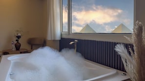 A bathroom with a bathtub filled with bubbles, a window with a view of pyramids, a radiator, and a small table with a vase and a bottle. - Pyramids Palace View (Giza)