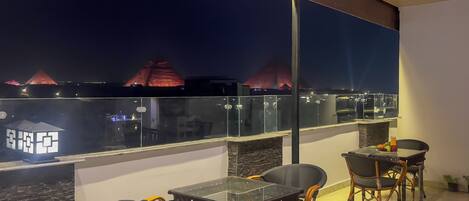 A balcony with a glass table and chairs, overlooking a cityscape with illuminated buildings.