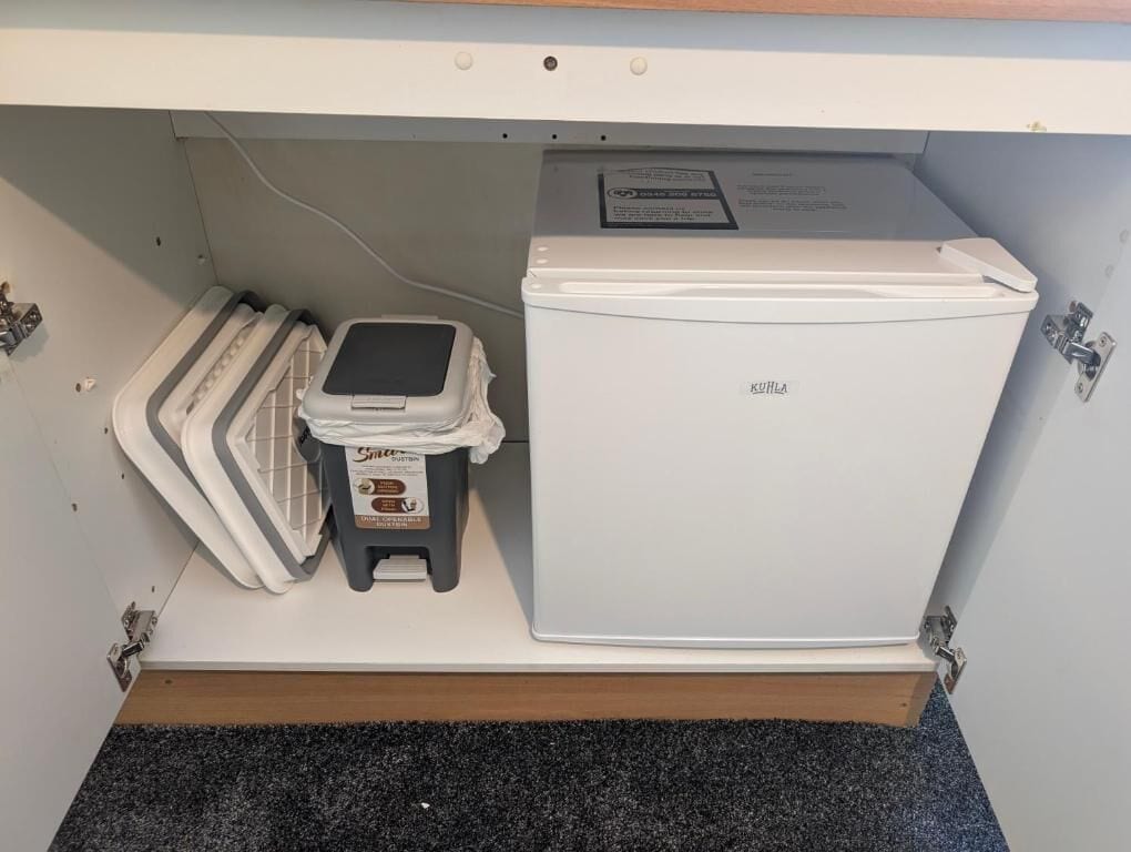 Een witte koelkast met een bedieningspaneel bovenop, een klein wit apparaat, en een opgevouwen mat op de vloer.