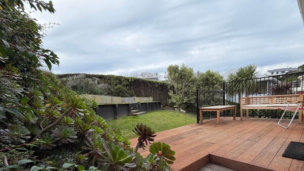 A wooden deck with steps leading to a seating area, surrounded by lush greenery and a view of a hill.