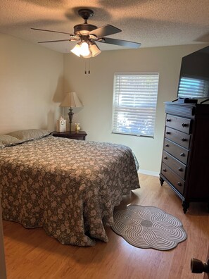 Une chambre à coucher comprenant un lit, une commode, un ventilateur de plafond, une lampe, une horloge, une fenêtre avec des stores et un téléviseur posé sur une commode.
