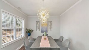 Een eetkamer met een rechthoekige tafel, grijze stoelen, een kroonluchter en kunstwerken aan de muren.