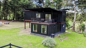 A modern two-story cabin with a balcony, surrounded by a grassy area and trees.
