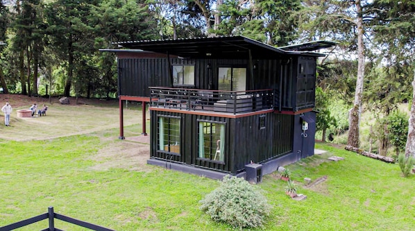 A modern two-story cabin with a balcony, surrounded by a grassy area and trees.