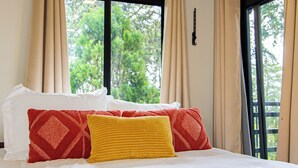 Quarto com teto de madeira, cama com roupa de cama branca e duas almofadas, janela com cortinas e vista para o verde lá fora.