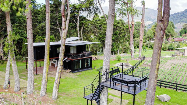 A modern house with a deck, surrounded by tall trees and a grassy area.
