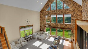 A spacious living area with a stone wall, large windows, a fireplace, and a staircase. - Mountaintop Cabin|Game Room|Pickleball (Elliottsburg)