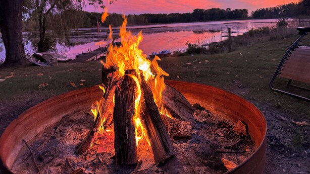 Un feu de camp dans un foyer métallique, entouré d’une zone herbeuse avec des arbres et un lac en arrière-plan.