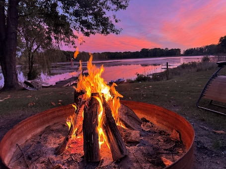A campfire in a metal fire pit surrounded by a grassy area with trees and a lake in the background.