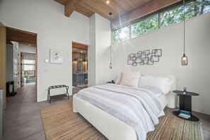 A modern bedroom with a large bed, a small side table, a hanging light fixture, and a view of the outdoors.